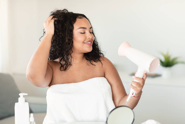 Női hajápolási koncepció. Afrikai amerikai test pozitív hölgy szárítás és stílus haj hajszárítóval, áll a hálószobában, törülközőbe csomagolva. Nő használ hajszárítót zuhanyzás után - Fotó, kép