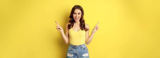 Portrait of stylish young female model, showing advertisement, pointing at logo or banner, demonstrating two ways, choices in store, standing over yellow background. - Zdjęcie, obraz