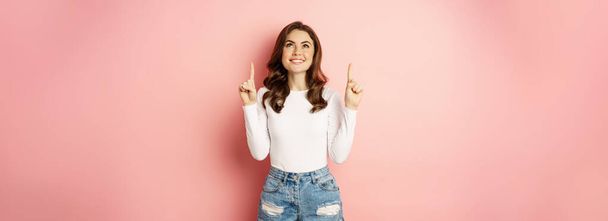 Pointing up. Happy smiling stylish girl, showing announcement on top, demonstrating advertisement, wearing stylish summer outfit, pink background. - Fotó, kép
