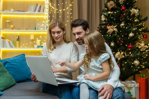 A child girl chooses a gift for Christmas in a laptop online, points to the monitor. Sitting with mom and dad on the sofa at home near the Christmas tree. - Valokuva, kuva