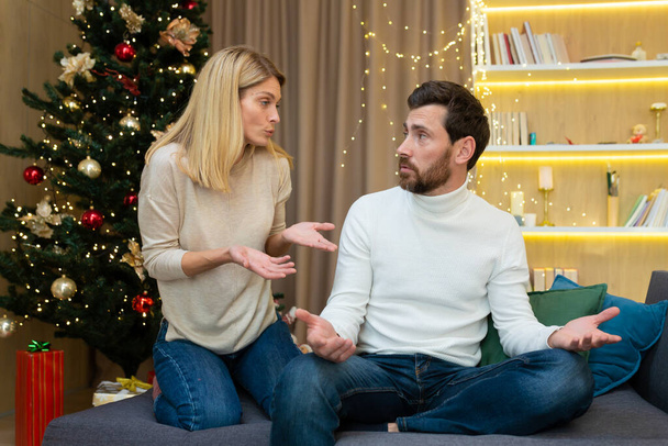 Family conflict on the holiday. A young couple, a family, a man and a woman quarrel at Christmas. They are sitting on the sofa at home near the Christmas tree, shouting at each other. - Foto, immagini