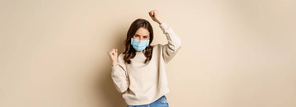 Covid-19, pandemic and quarantine concept. Enthusiastic girl dancing in face medical mask, celebrating, standing over beige background. - Zdjęcie, obraz