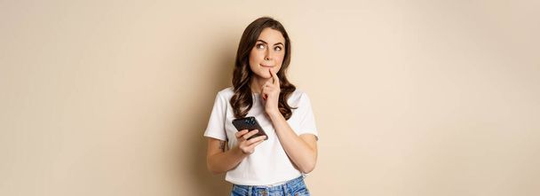 Girl thinks, holds mobile phone, looks up thoughtful, stands over beige background. - Foto, Imagen