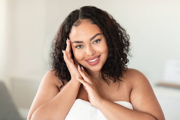 Ritratto di bella donna afroamericana paffuta che tocca il viso mentre posa avvolta in un asciugamano e sorride alla macchina fotografica. Il concetto di bellezza, benessere e spa - Foto, immagini