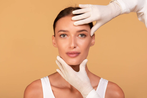 Cosmetische chirurgie concept. Dokter onderzoekt jonge Kaukasische vrouwen gezicht over beige studio achtergrond. Portret van een mooie dame tijdens een bezoek aan het kosmetoloog kabinet - Foto, afbeelding
