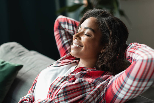 Glad happy pretty black young female with closed eyes resting alone on sofa, enjoys silence and comfort in minimalist living room interior, close up. Break, relax in cozy home, lifestyle at weekend - Фото, зображення