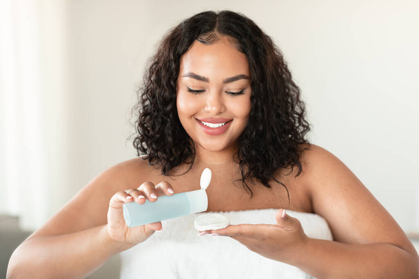 Привлекательное черное тело позитивная женщина, использующая хлопчатобумажные прокладки и мицеллярную воду для ухода за кожей, очищая кожу, сидя в спальне интерьера дома - Фото, изображение