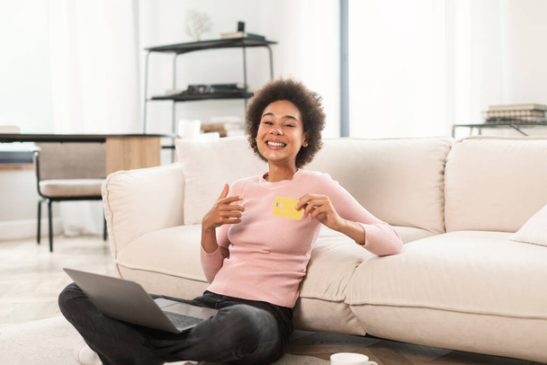 Glad millennial african american woman with credit card, laptop shopping online, sitting on floor in living room interior. Huge sale, banking finance and money management, cashback at home, covid-19 - Fotografie, Obrázek