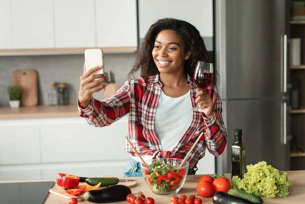 Sonriente mujer joven bonita negro en informal con ensalada y copa de vino hace selfie o tiene videollamada en el interior de la cocina moderna con verduras orgánicas. Reunión remota, aplicación para el blog de alimentos en casa - Foto, Imagen