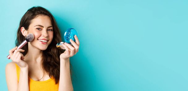 Beauty. Close up of attractive woman looking in mirror and using brush to apply make up, smiling pleased at camera, standing against blue background. - Photo, Image