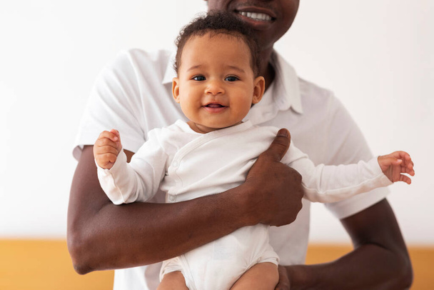 Concepto de cuidado infantil. Padre sosteniendo lindo bebé negro en las manos, sonriendo joven afroamericano abrazando a su adorable hijito o hija, niño pequeño mirando a la cámara, primer plano - Foto, Imagen