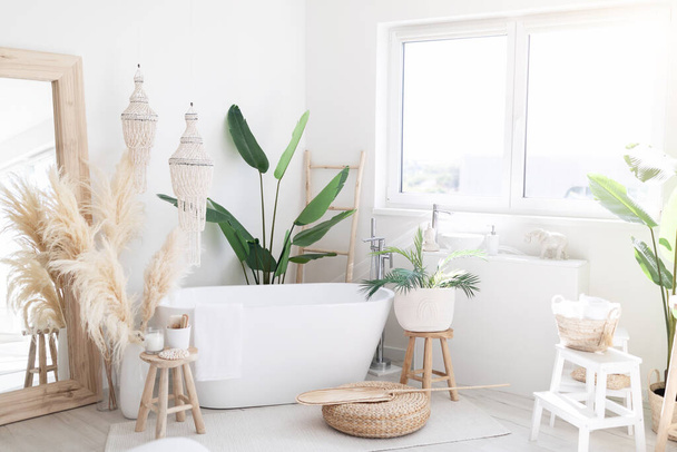 Стильний інтер'єр світлої ванної кімнати з русичними прикрасами в стилі бохо, затишна простора ванна кімната з натуральними зеленими рослинами біля вікна та великою білою ванною всередині, вільний простір
 - Фото, зображення