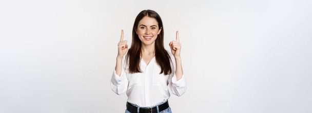 Smiling confident pretty woman, pointing fingers up, showing logo banner, announcement, standing in white blouse against studio background. - Foto, Imagen