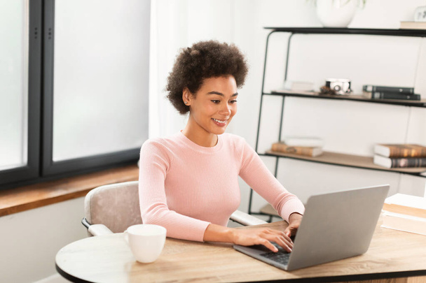 Cheerful millennial african american businesswoman typing on laptop, sitting at table in bright office interior, free space. Business remote, work, freelance, video call and social distance at home - Foto, Imagem