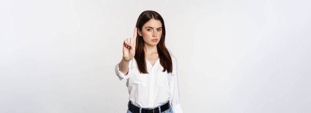 Serieuze vrouw toont taboe, stop gebaar, schudt vinger in afkeuring, oneens, verbiedt iets, staat over witte achtergrond. - Foto, afbeelding