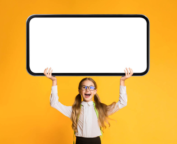 Podekscytowany Little Schoolgirl Holding Big Blank Smartphone z białym ekranem nad głową, Zaskoczony nastolatek Kobieta Reklama edukacyjna App or Website, Stojąc na żółtym tle, Mockup - Zdjęcie, obraz
