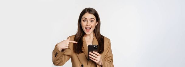 Venditrice appassionata, donna d'affari che punta il dito sul cellulare e sorride, mostra sul cellulare, in piedi su sfondo bianco. - Foto, immagini
