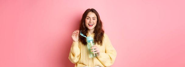 Portrait of happy brunette woman with curly hairdo, drinking water with lemon from personal bottle, laughing and smiling, feeling healthy, standing against pink background. - Foto, Imagem