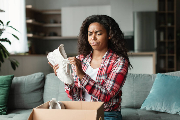 Sad displeased angry black young woman in casual unpacks parcel, looks at new sneakers in living room interior. Shopaholic emotions from online shopping, mistake or damaged purchase and delivery order - Photo, image