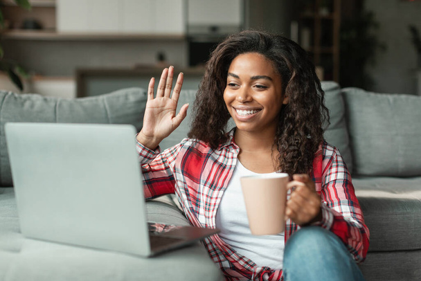 Cheerful pretty black young woman in casual with cup of hot drink waving her hand at laptop webcam in living room interior. Social networks, video call, meeting remotely, online chat and coffee break - Photo, Image