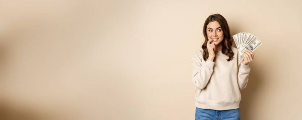 Excited brunette woman holding money and biting finger with yearning face, watns to buy smth, shopoholic with cash, standing over beige background. - 写真・画像