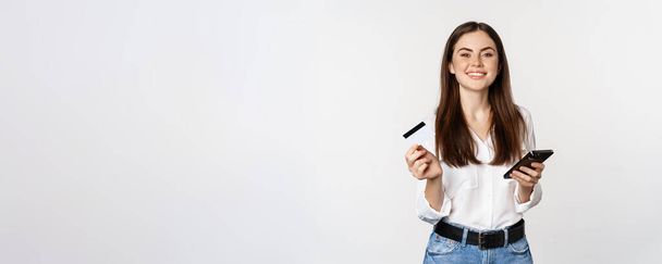 Sonriendo mujer moderna usando tarjeta de crédito y teléfono móvil, pagando, haciendo la compra en línea, compras en el teléfono inteligente, de pie sobre fondo blanco. - Foto, imagen