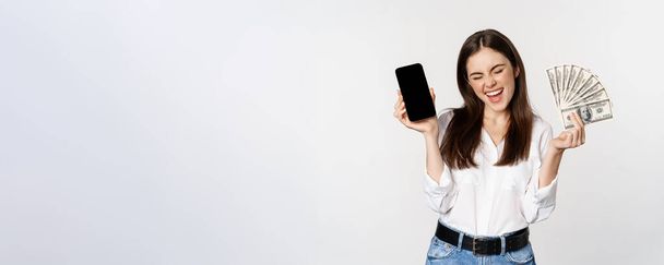 Enthusiastische junge Frau gewinnt Geld, zeigt Smartphone-App-Schnittstelle und Bargeld, Mikrokredite, Preiskonzept, steht vor weißem Hintergrund. - Foto, Bild