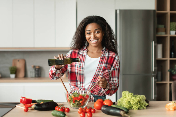 Smiling glad pretty black young female chef in casual pours oil at salad from organic vegetables in minimalist kitchen interior, copy space. Cooking food at home, diet, healthy homemade dinner alone - Photo, Image