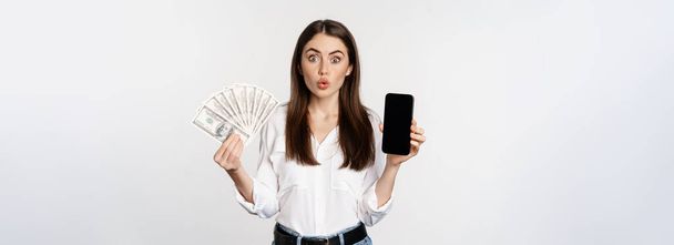 Ενθουσιώδης νεαρή γυναίκα κερδίζει χρήματα, δείχνει διεπαφή εφαρμογή smartphone και μετρητά, microcredit, βραβείο έννοια, στέκεται πάνω από το λευκό φόντο. - Φωτογραφία, εικόνα