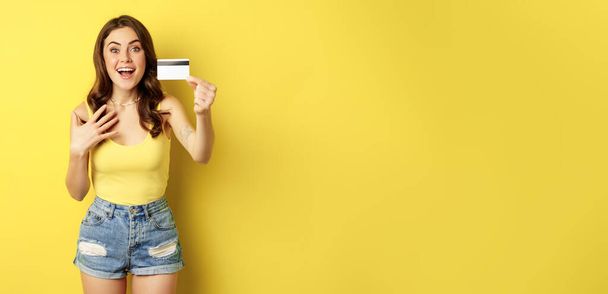 Porträt eines brünetten Mädchens, das seine Kreditkarte zeigt und lächelt, Sommerkleidung trägt, Konzept der Bank, Geld und Einkaufen, vor gelbem Hintergrund stehend. - Foto, Bild