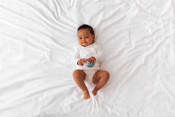 Top View Of Charming Little Black Baby With Rattle Toy In Hands Relaxing In Bed At Home, Above Shot Of Cute Adorable Infant African American Boy Or Girl Resting In Bedroom, Copy Space - Valokuva, kuva