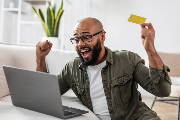Online winkelen. Vreugdevolle Latijnse man met laptop en creditcard, gebaren ja tijdens het maken van een succesvolle betaling, zitten in de woonkamer interieur thuis - Foto, afbeelding