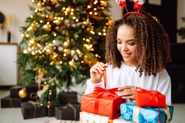 Mladá usmívající se krásná brunetka žena s dárkovou krabicí ve zdobeném interiéru s vánoční stromeček. Nový rok.  - Fotografie, Obrázek