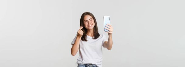 Mutlu, güzel, genç bir kadın kalp hareketi yapıyor, akıllı telefondan selfie çekiyor ve gülümsüyor.. - Fotoğraf, Görsel