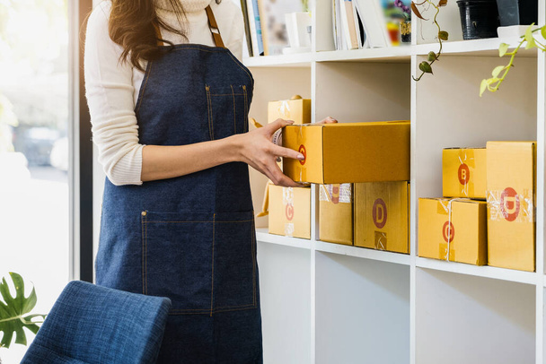 家から仕事だ。オンラインで製品を販売する幸せな女性スタートアップの小さなビジネスの所有者は、顧客の注文をパックし、郵便料金のためにそれらを準備するために小包ボックスを拾う - 写真・画像