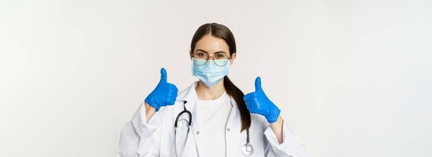 Ritratto ravvicinato di medico donna, medico in maschera facciale da coronavirus, mostrando i pollici in alto e sorridente, raccomandando clinica o sito web medico, sfondo bianco. - Foto, immagini
