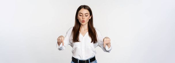Surprsed businesswoman, beautiful adult woman pointing fingers down, looking excited at camera, standing over white background. - Фото, изображение