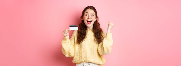 Excited young woman winning prize, see awesome promo sale, showing her plastic credit card and scream of joy, saying yes, standing against pink background. - 写真・画像