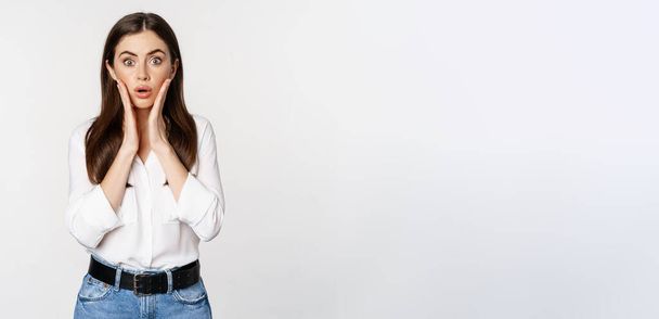 Portrait of concerned female model looking shocked, surprised face reaction, standing in shirt over white background. Copy space - Photo, image