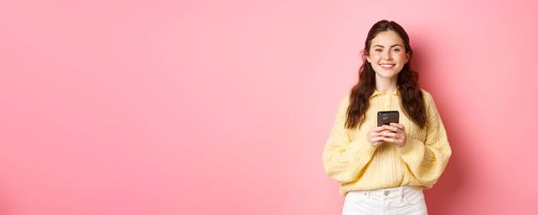 Портрет красивой девушки с мобильным телефоном и улыбкой, покупки в Интернете, с помощью приложения социальных сетей, стоя на розовом фоне в повседневной одежде. - Фото, изображение