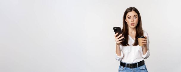 Portret młodej kobiety wyglądającej na zaskoczoną po sprawdzeniu telefonu komórkowego, trzymającej filiżankę kawy na wynos, stojącej nad białym tłem. - Zdjęcie, obraz