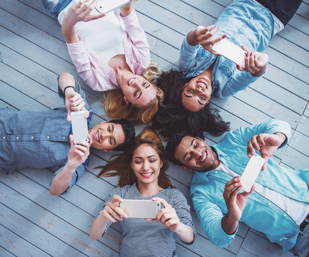 Вид сверху на молодых людей разных национальностей, которые пользуются смартфонами и улыбаются, лежа на полу
 - Фото, изображение