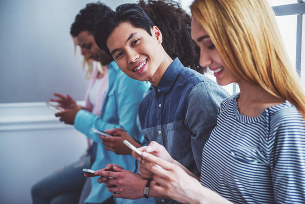 Young people of different nationalities using smartphones and smiling while sitting in a row. Handsome Asian guy is looking at camera - Photo, Image