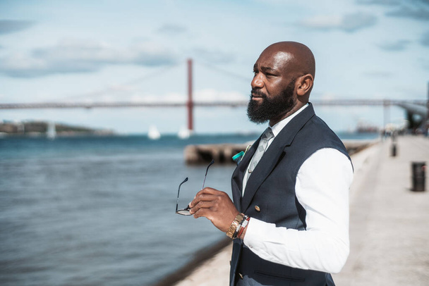 Portrait of a mature handsome bald African American businessman with a well-groomed black beard and in a tailored formal suit with a vest, holding his eyeglasses in the hand while standing on the pier - Фото, зображення