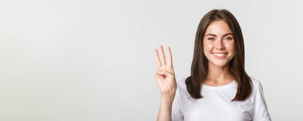 Κοντινό πλάνο χαρούμενα ελκυστική νεαρή γυναίκα χαμογελώντας, δείχνει τρία δάχτυλα, λευκό φόντο. - Φωτογραφία, εικόνα