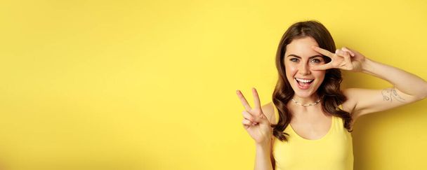 Close up portrait of stylish happy girl, showing peace sign and smiling, posing against yellow background. Copy space - Photo, Image