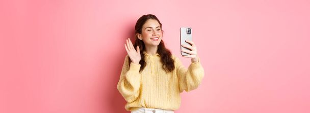 Portrait of friendly girl talking on video chat app, waving hand at smartphone camera, having mobile conversation, standing against pink background. - Photo, Image