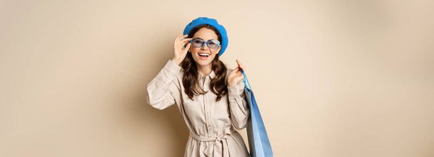 Élégante fille heureuse sur le shopping. Portrait de femme moderne avec sac de magasin, riant et souriant satisfait, s'achetant un cadeau, debout sur fond beige. - Photo, image