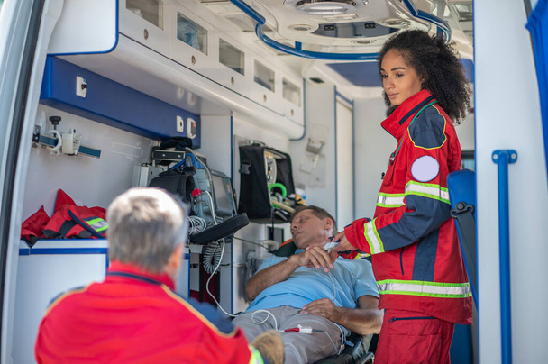 Poważny lekarz ambulansu gapiący się na jej kolegę podczas pomiaru poziomu tlenu we krwi nieprzytomnego mężczyzny - Zdjęcie, obraz