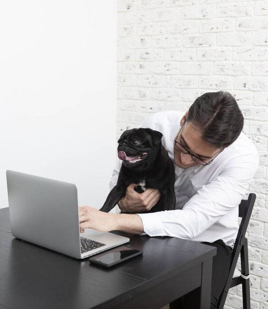 Hombre de negocios sonriente y su encantador pug mascota trabajando en el ordenador portátil en casa. Conceptos de trabajo distante y amistad - Foto, imagen